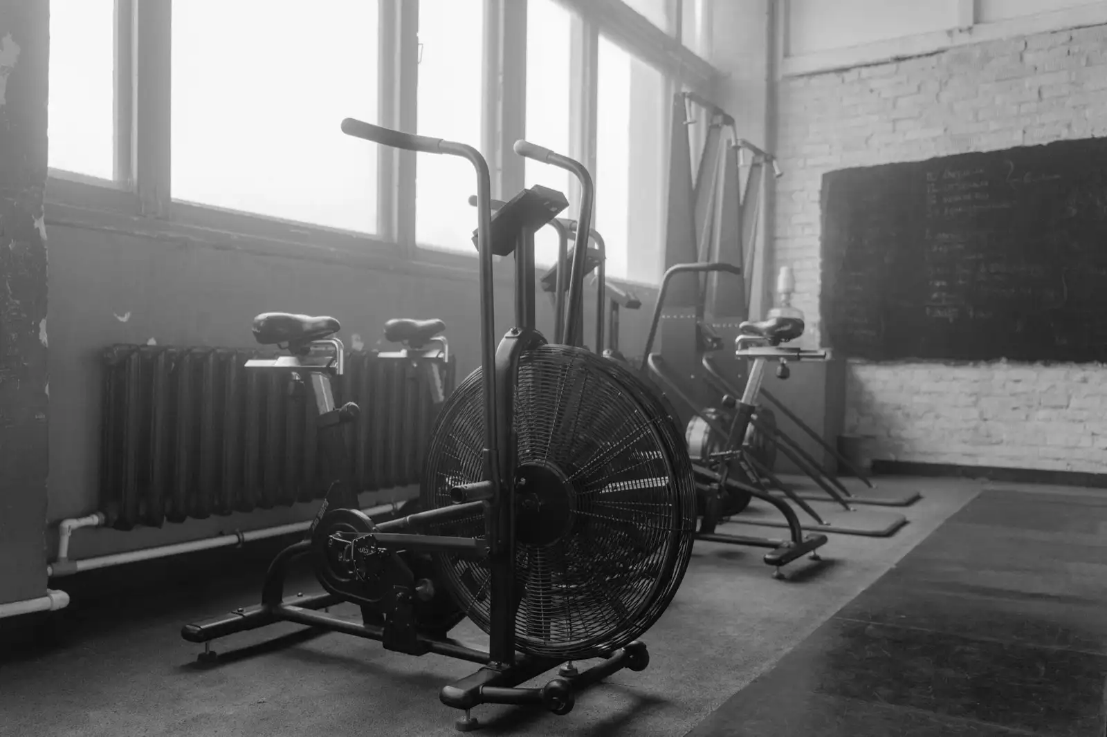 Exercise bikes lined up by tall windows in a gym, black and white